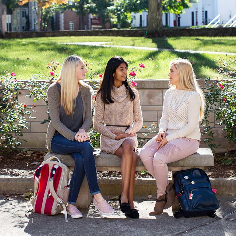 Three female students sit on a bench in front of rose bushes on the IU Bloomington campus.